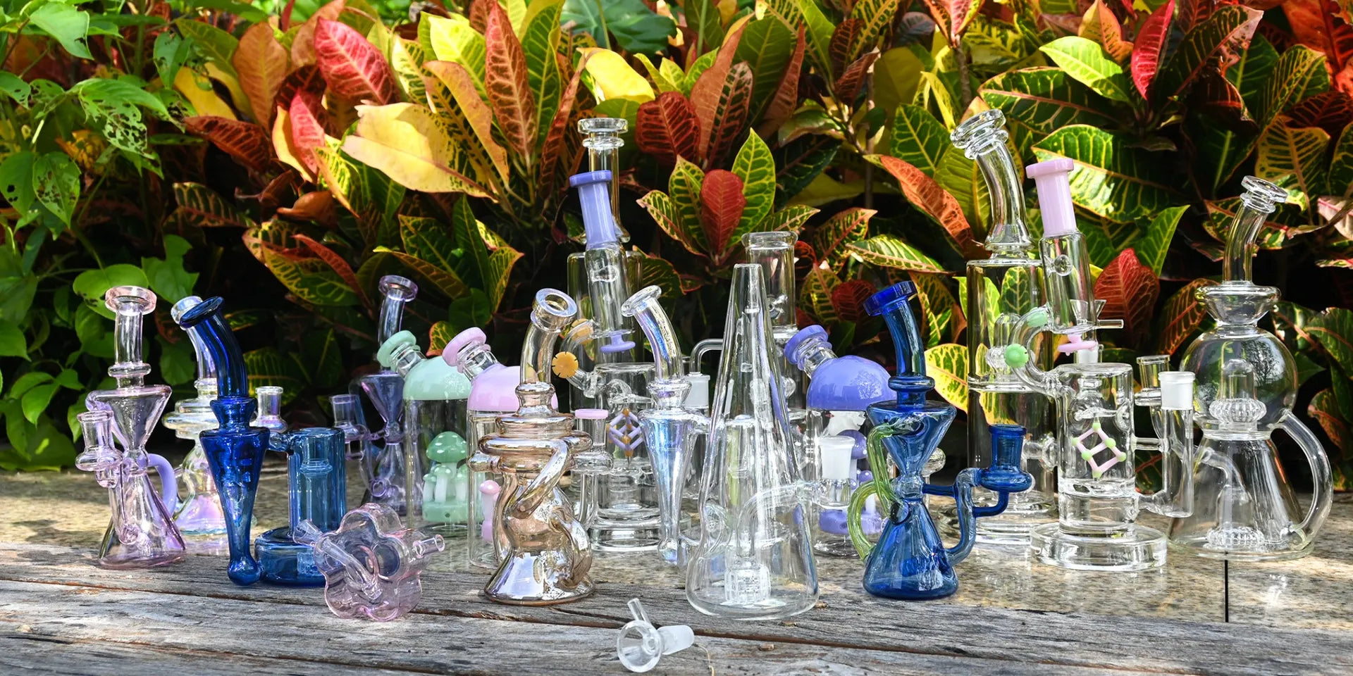A diverse collection of premium borosilicate glass bongs, recycler dab rigs, and mini rigs from Croia Glass, displayed outdoors on a wooden table with lush tropical foliage.